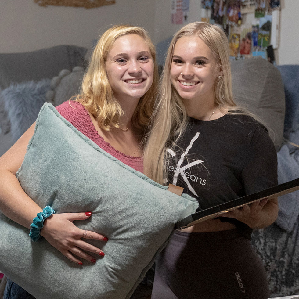 Two Skidmore students smiling together in a cozy residence hall room, holding a pillow and picture frame during move-in.