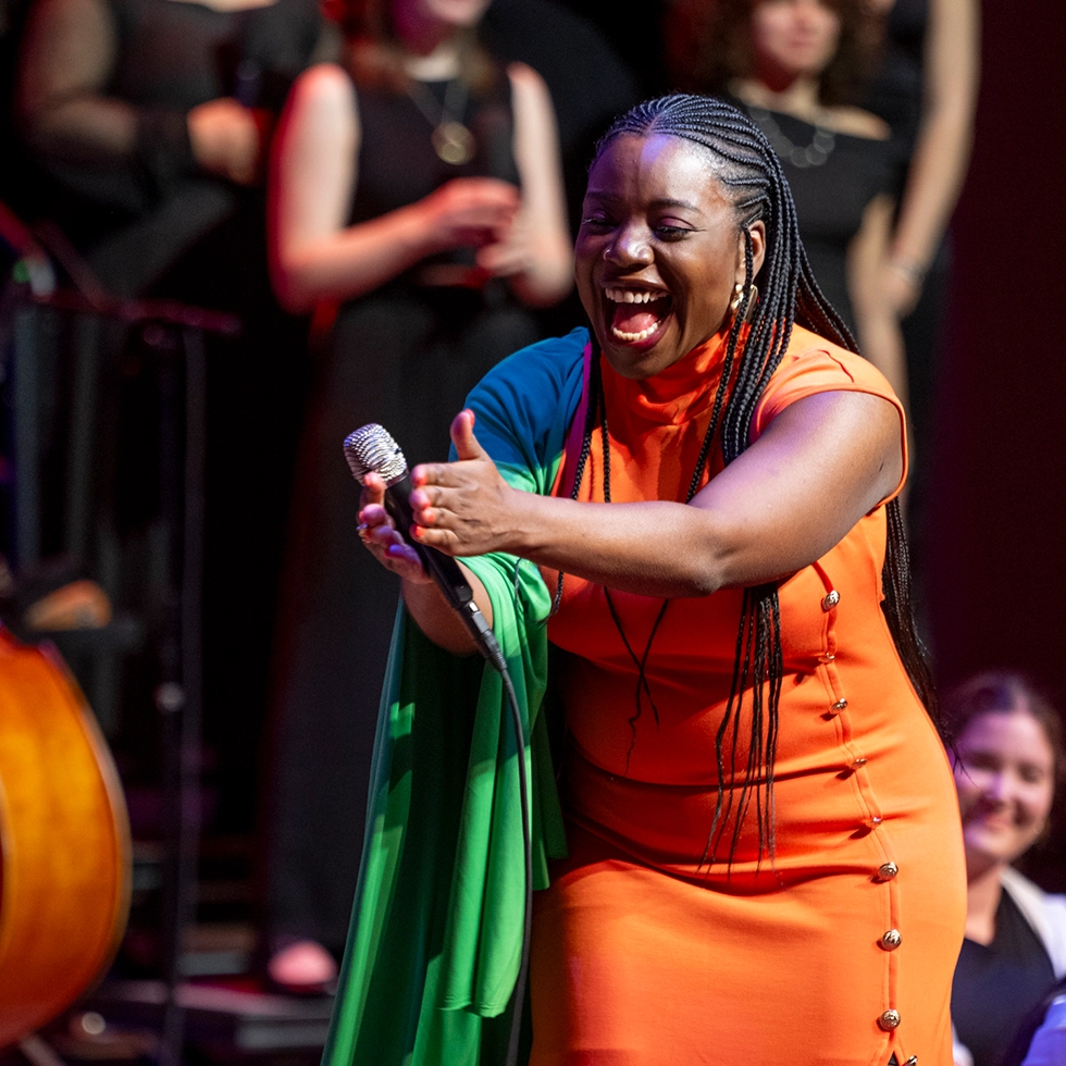 A performer in an orange dress and green shawl passionately sings and gestures on stage during a musical performance at Skidmore College.