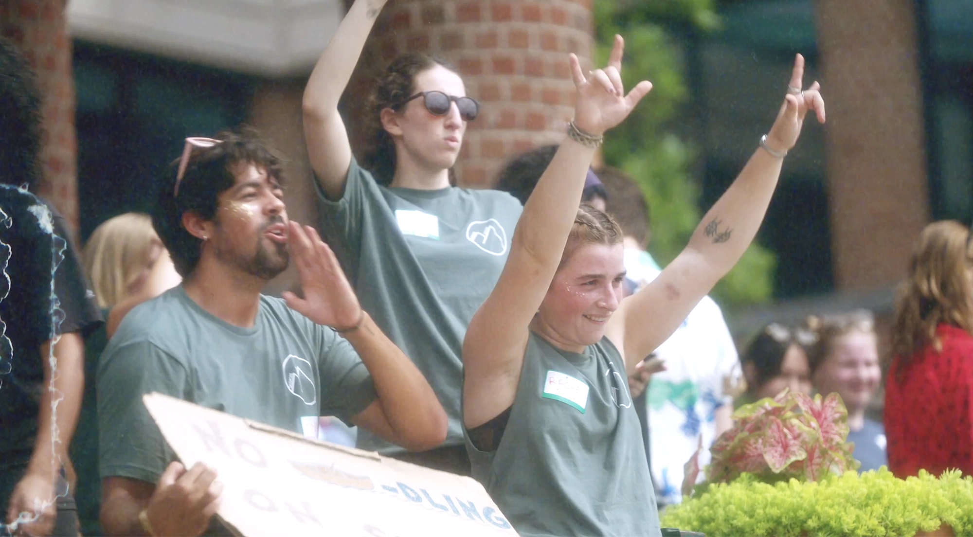 Group of Skidmore College students enthusiastically cheering and raising their arms during a campus event. They wear matching green t-shirts with name tags and a Skidmore logo.