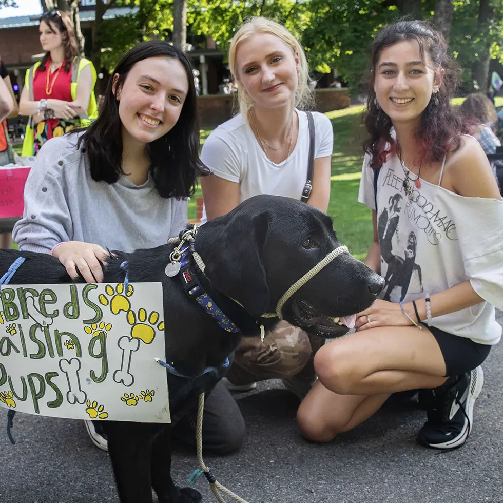 Three students kneel next to a black lab wearing a “T-Breds Raising Pups” sign at Skidmore’s student club fair.