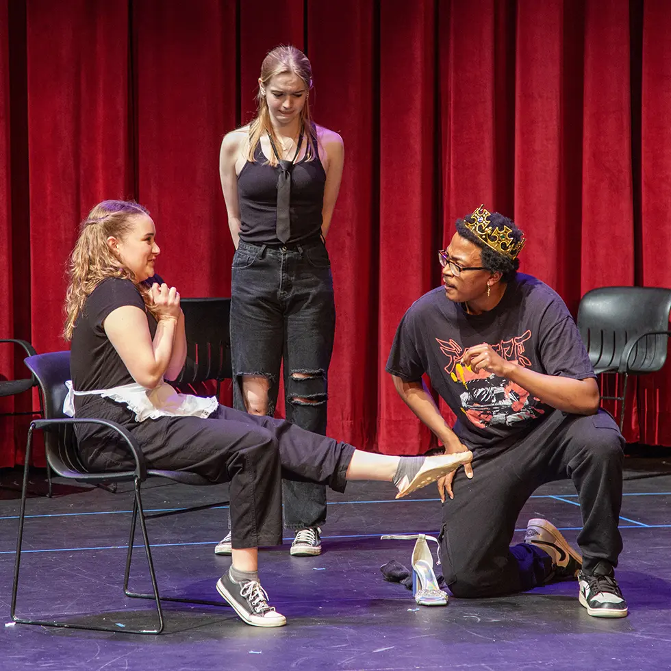 Three Skidmore students perform a humorous theater scene onstage, with one pretending to fit a glass slipper in front of a red curtain.