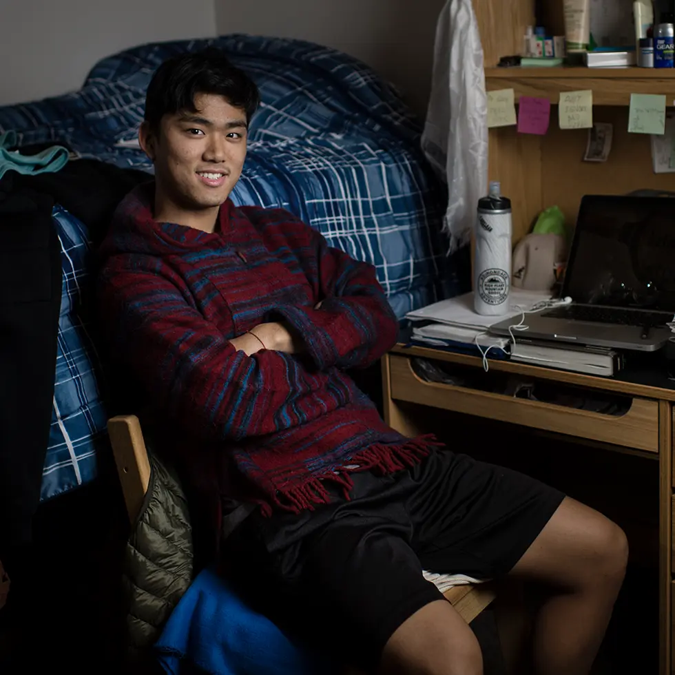 A Skidmore student sits comfortably in a residence hall room, smiling with arms crossed beside a neatly made bed and a personalized desk filled with study materials and personal items.