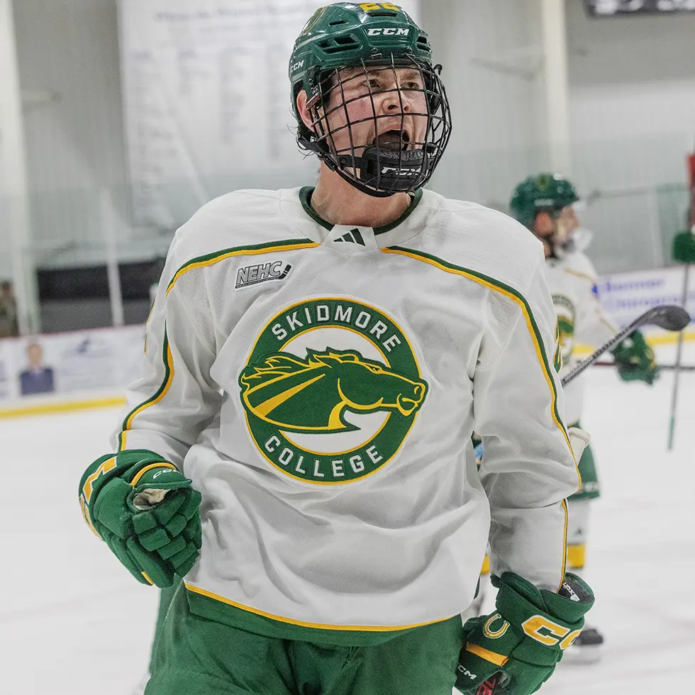 A Skidmore hockey player cheers on the ice in front of a crowd during the Pack the Rink event, wearing the Thoroughbred team jersey.