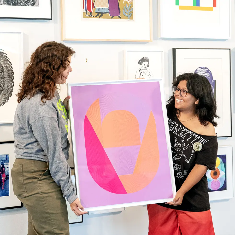 Two students smile while holding up a framed abstract print in Skidmore’s on-campus gallery, surrounded by colorful artwork.