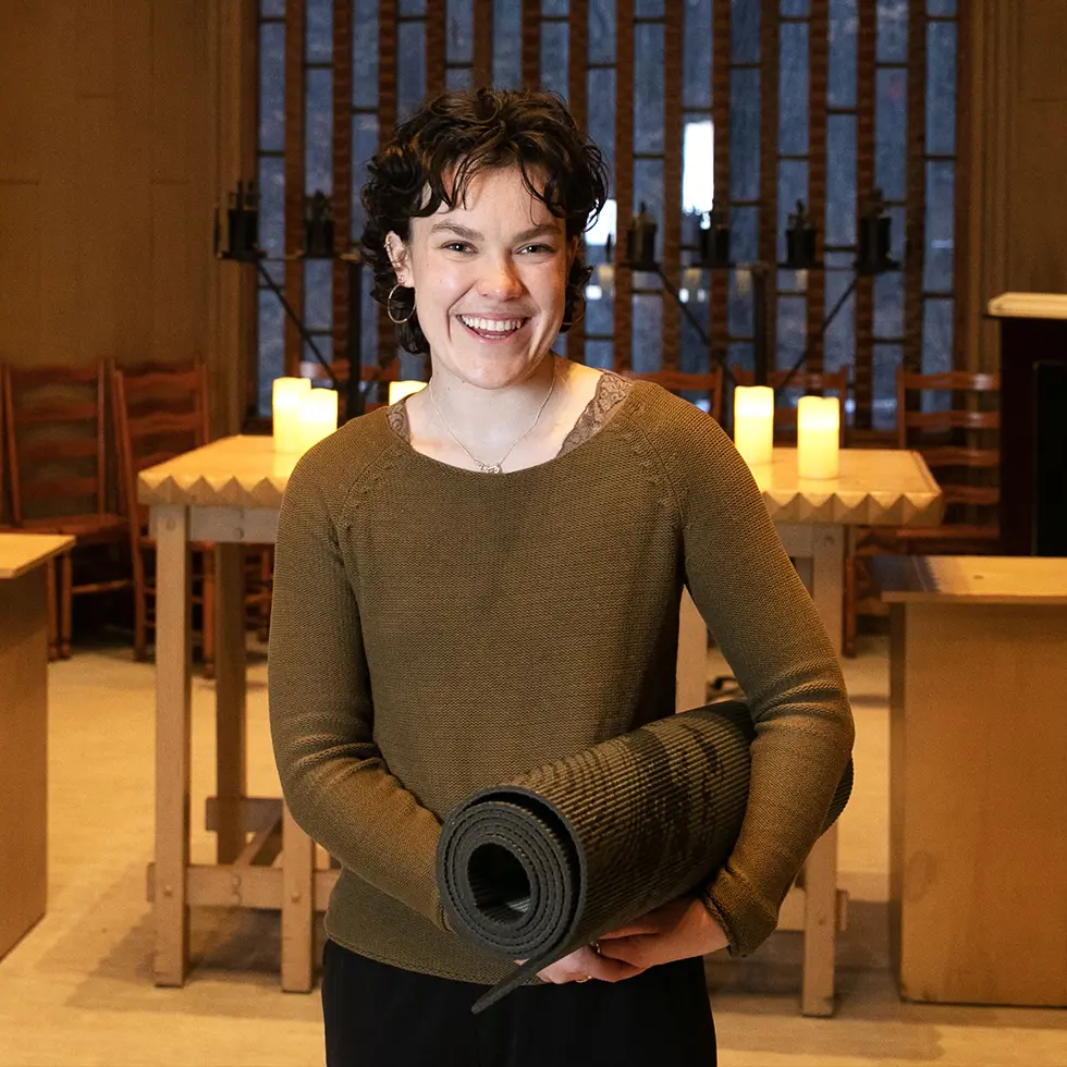 A student stands in the Skidmore campus chapel holding a rolled yoga mat, surrounded by wooden furniture and soft candlelight.