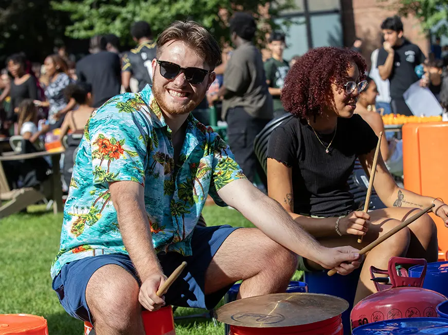 Students enjoy an outdoor drum circle during a sunny afternoon event, smiling and playing instruments in a lively crowd.