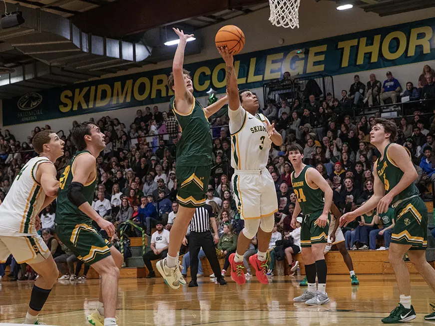 A Skidmore student-athlete leaps toward the hoop mid-game in front of a packed crowd, surrounded by opposing players in green uniforms.