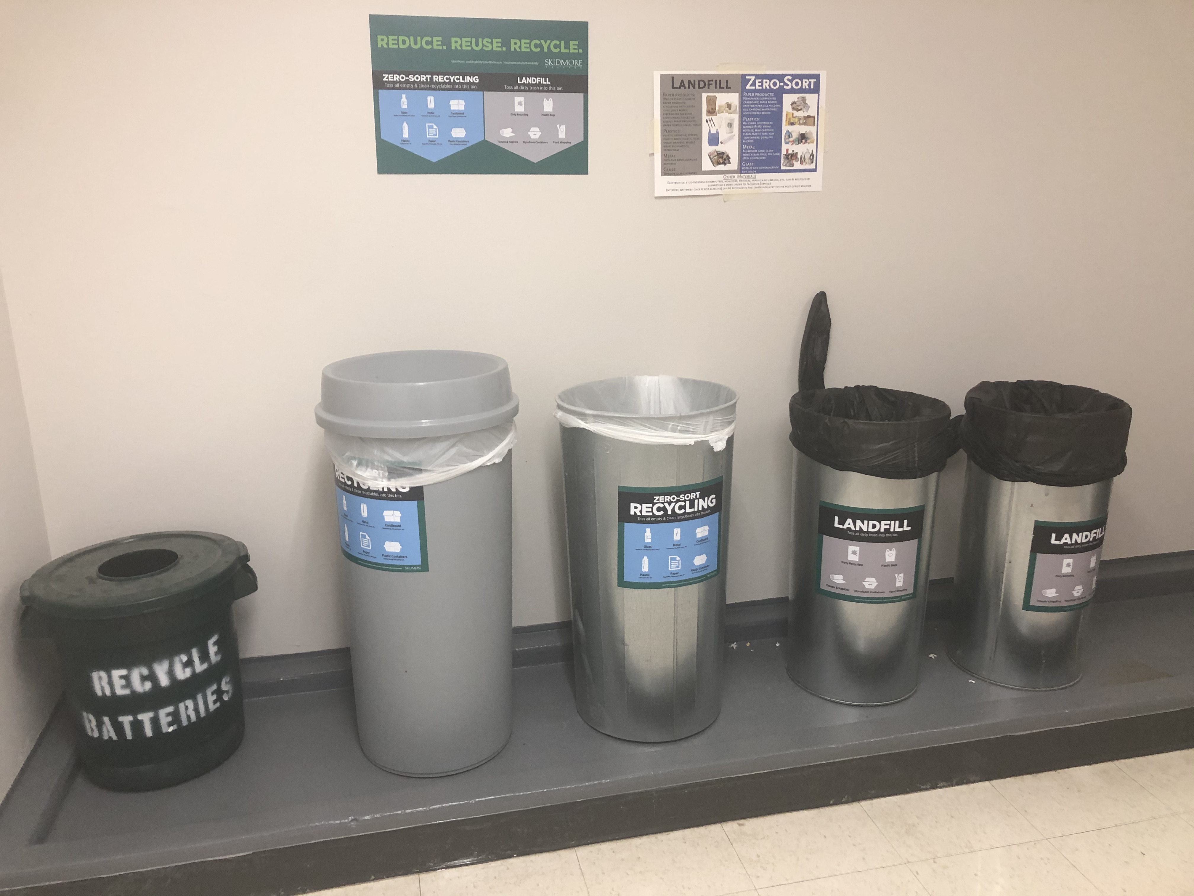 5 bins lined up next to each other in a hall kitchen space. 1 gray bin for batteries, 2 tall bins labelled recycling and 2 tall bins labelled landfill.