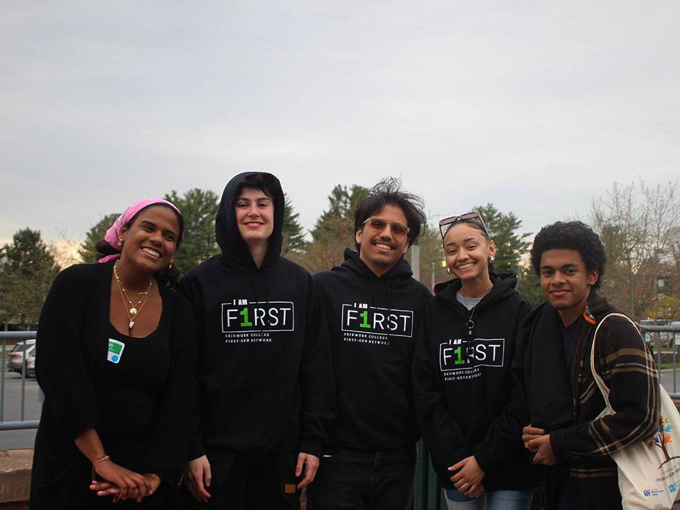 Five students stand outside posing in a line together, wearing black Skidmore first-generation student shirts 