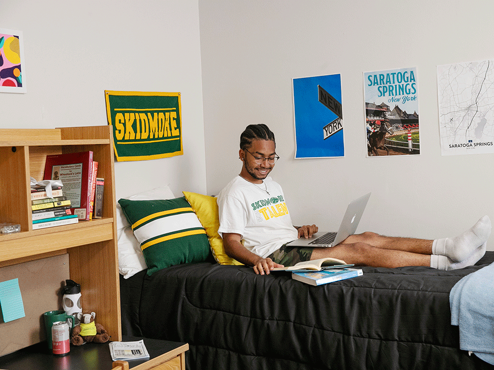 Student sits on a dorm bed with his laptop on his lap and books at his side.
