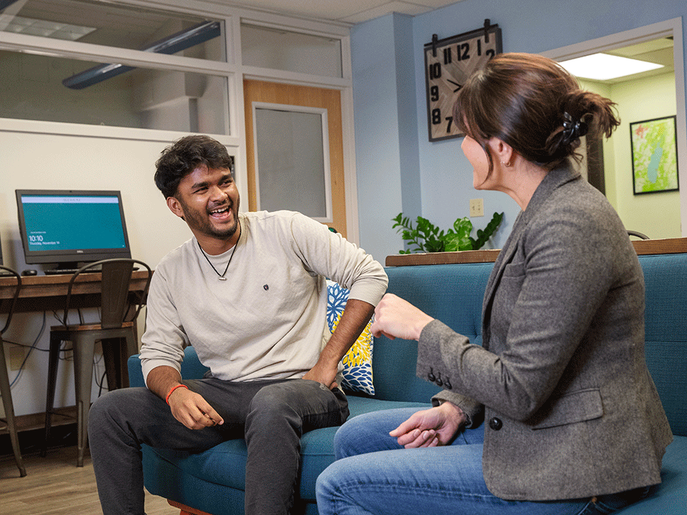 A student and an academic counselor sit on a couch laughing during a discussion.