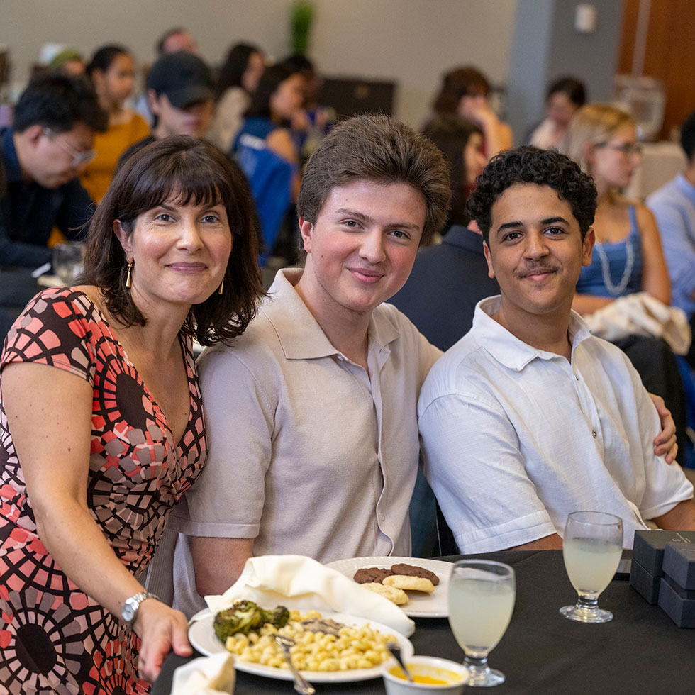 Two students sit at a table posing with Chloe Jaleel with a crowded room of people sitting in the background