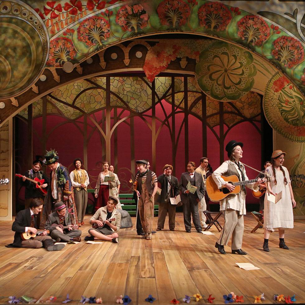 A large group of students perform on stage to 'As you like it'. Some hold instruments, some are sitting on the ground, while others are standing. All are singing with an intricate set design in the background.