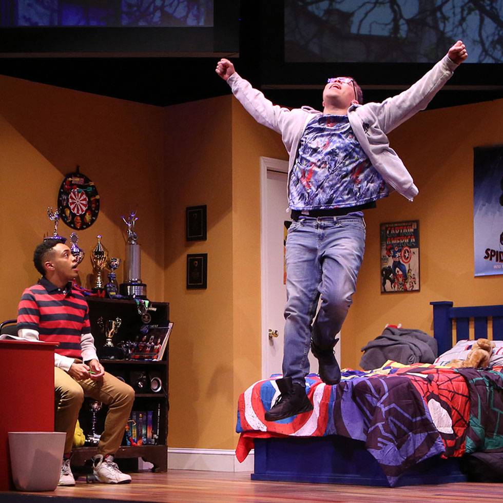 Two theater students perform on stage set up as a bedroom. One actor is jumping off the bed with their arms raised out, while the other sits in a chair watching.