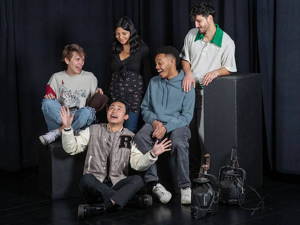 Miranda fellows sit together on a black stage making playful faces at each other and smiling. The one sitting in front of the other four has their arms raised out and hands opened wide.