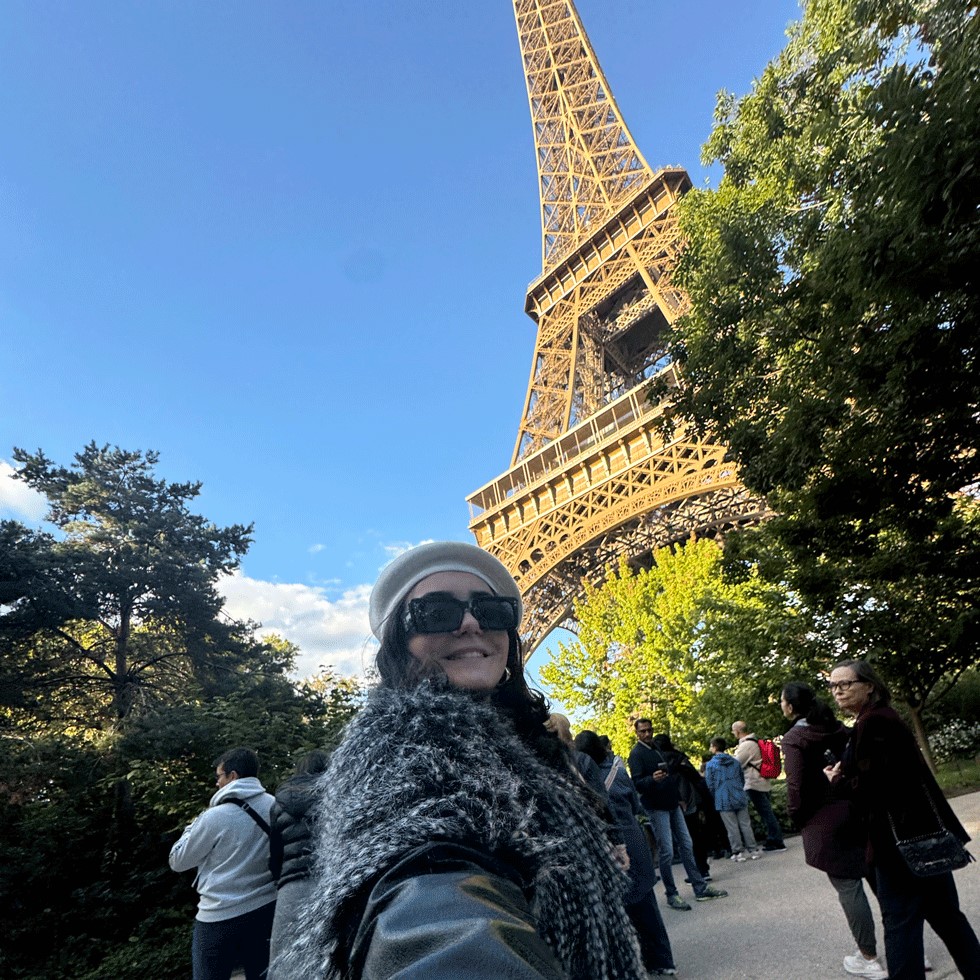 Student takes selfie with Eiffel Tower in the background on a sunny blue sky day. They smile while wearing a beret and sunglasses.
