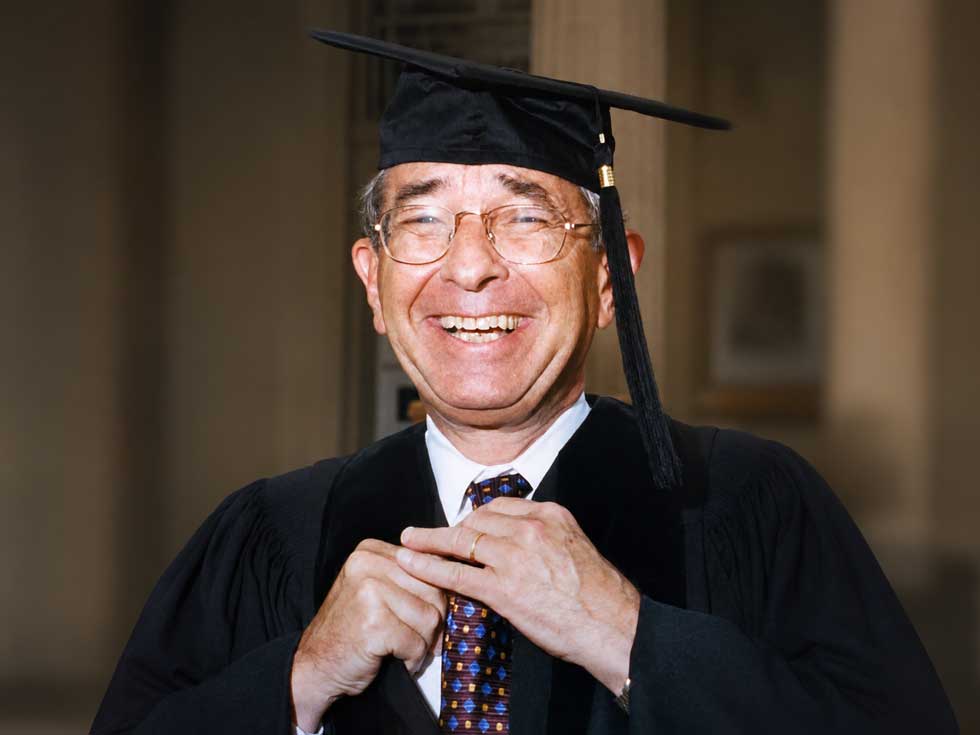 Arthur Zankel smiles in academic regalia as he adjusts his tie while wearing a mortarboard cap.