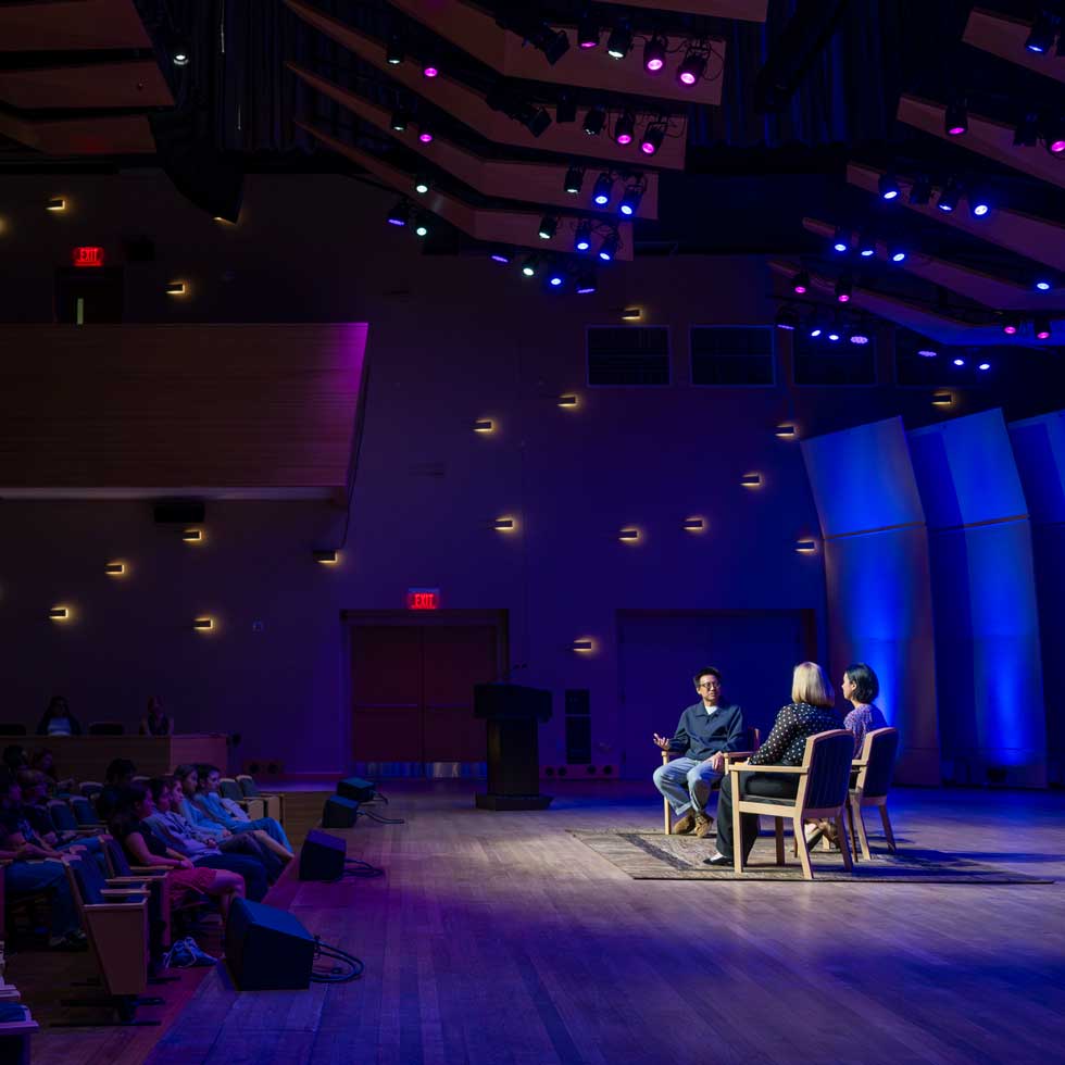Speakers participate in a seated conversation onstage in Zankel Music Center while an audience listens under blue and purple stage lighting.