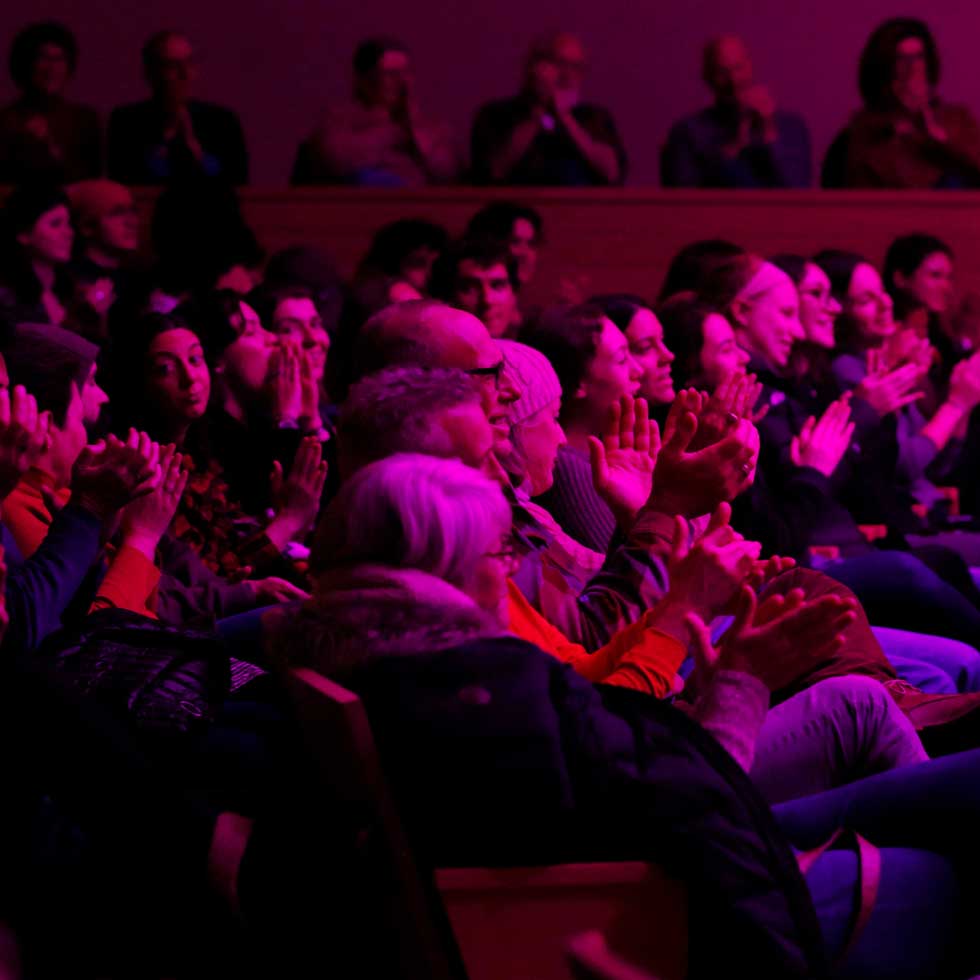 Audience members applaud during a performance inside Zankel Music Center under purple and pink concert lighting.