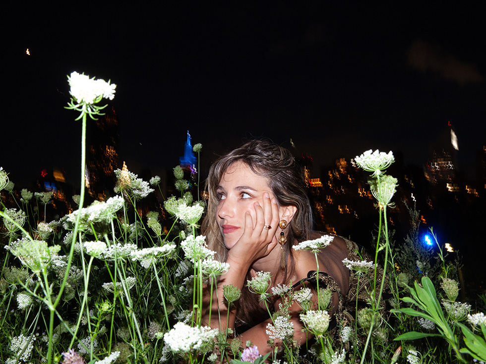 A woman rests her chin on her hand among white wildflowers at night, with city lights glowing in the background.