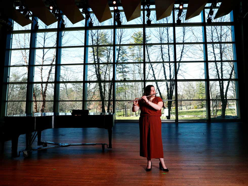 A flutist performs onstage inside Zankel Music Center, with a grand piano and tall windows behind her.