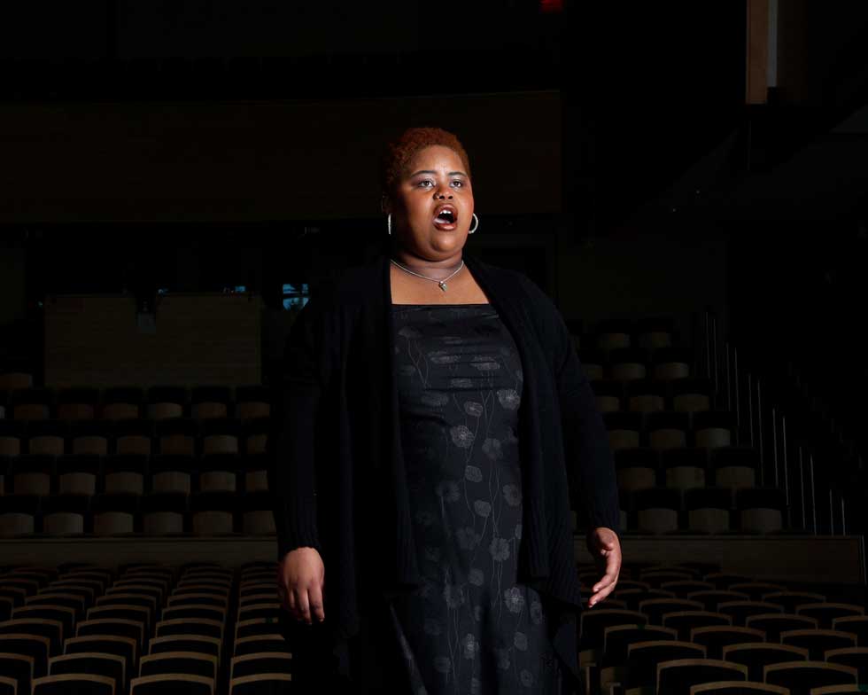 A vocalist sings onstage in a darkened concert hall with rows of seats behind her.