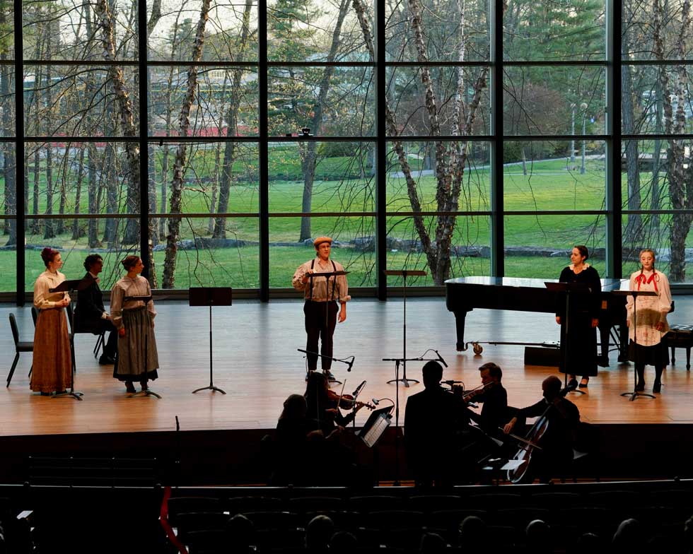 Vocalists and musicians perform on Zankel Music Center’s stage in front of tall windows overlooking campus trees.